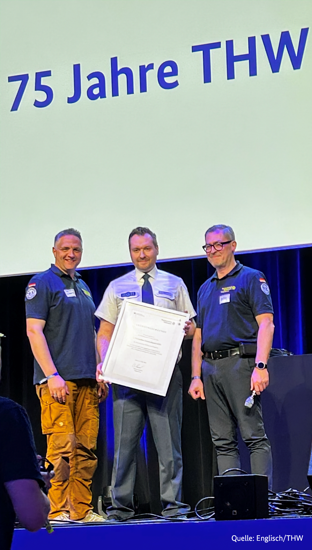 Man sieht 3 Männer auf einer Bühne, die eine Urkunde halten und glücklich aussehen, im Hintergrund steht: 75 Jahre THW.
